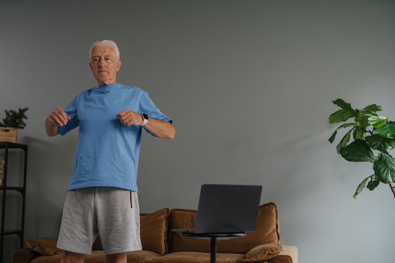 Elderly man doing exercise at home in front of a laptop, wearing a smartwatch.